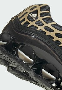 Närbild av en svart och guldfärgad Adidas-sneaker med fjäderbelastade stötdämpande element i sulan för stötupptagning och stabilitet.