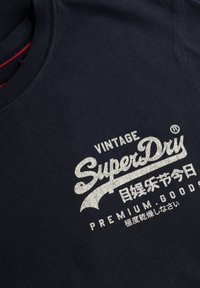 Donkerblauw shirt met witte tekst "Vintage SuperDry Premium Goods" en Japanse karakters gedrukt op de linkerborst aan de voorkant.