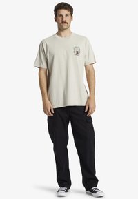 Camiseta de algodón beige con mangas cortas y un bolsillo en el pecho que presenta un diseño gráfico colorido. Combinada con pantalones cargo negros y zapatillas.