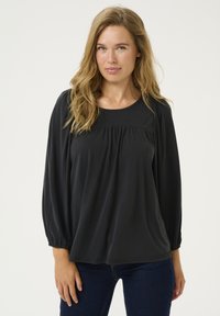 Črna bluza z dolgimi rokavi, okroglim izrezom, zgibanim sprednjim delom in nabranimi manšetami, narejena iz mehkega materiala. V kombinaciji s temnimi jeans hlačami.