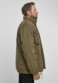 Olive green military-style jacket made of cotton fabric. Features a zip and snap closure, multiple pockets, and decorative stitching details.