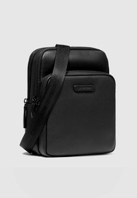 Schwarze Crossbody-Tasche aus Leder mit strukturierter Oberfläche, ausgestattet mit zwei Reißverschlüssen, einem breiten verstellbaren Riemen und einem Logo-Akzent auf der Vorderseite.