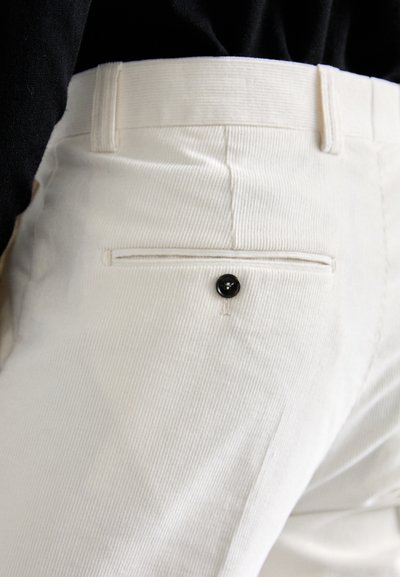 Detalhe em primeiro plano de um bolso de calças brancas com botão preto, usado com uma blusa preta.