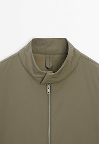 Veste vert olive avec col montant et fermeture éclair argentée visible, vue de face avec un focus sur la partie supérieure de la poitrine et le col.