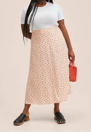 Jupe midi fleurie couleur pêche clair avec imprimé marguerites ; associée à un haut blanc ajusté et des sabots noirs ; tenant un petit sac tissé rouge.