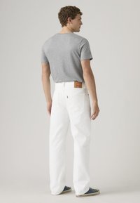 Νεαρός άνδρας που στέκεται πλάτη, φοράει γκρι μπλουζάκι, άσπρο τζιν με δερμάτινη ετικέτα και μπλε αθλητικά παπούτσια σε απλό φόντο.