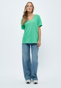 Žalias V tipo marškinėliai su trumpomis rankovėmis, derinami su šviesiai mėlynomis plačiomis kelnėmis, apsirengti su kasdieniškais pilkais sportbačiais, neutralioje fone.