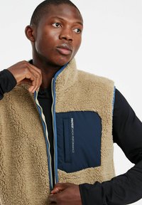 Beige Fleece-Weste mit einer dunkelblauen Brusttasche und Reißverschluss. Besitzt einen hohen Kragen und kontrastierende Abschlüsse. Weicher Stoff und lässiges Design.