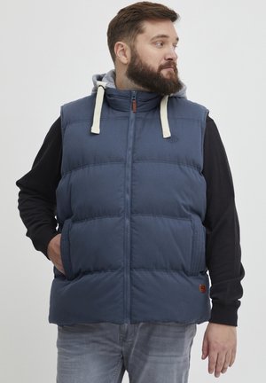 Tamno plavi puffer prsluk s visokim vratom, prednjim zipom i kremastim vezicama, nošen preko crne košulje dugih rukava. Ima bočne džepove.
