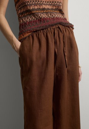 Nahaufnahme einer Person, die braune Leinen-Hose mit Kordelzug und ein mehrfarbiges gestricktes ärmelloses Oberteil trägt, mit den Händen in den Taschen.