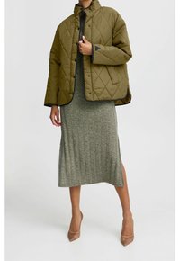 Giacca verde oliva trapuntata con colletto alto su un vestito grigio a coste, con uno spacco e scarpe con tacco neutre. Design semplice e pulito.