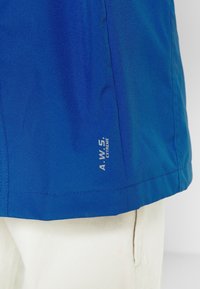 Ourlet d'une veste d'extérieur bleue avec le texte « A.W. 5. EXTREME » sur un pantalon blanc vu de côté.