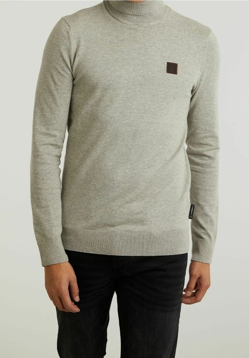 Homme portant un pull gris clair à côtes avec col rond et petit logo carré marron sur la poitrine, ainsi qu'un jean noir, debout devant un fond uni.