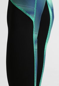 Leggings noirs avec un motif géométrique dynamique vert et bleu sur les côtés, présentant un tissu texturé et des coutures contrastées.