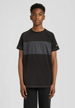 Dreng står med front mod kamera iført en sort t-shirt med en mørkegrå horisontal stribe og sorte bukser mod en ensartet lys baggrund.