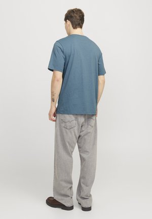 Man standing facing away wearing a blue short-sleeve T-shirt, light gray loose jeans, and dark shoes on a plain background.