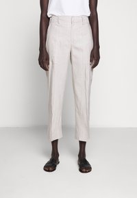 Beige linnebyxor med hög midja, sidofickor och smalnade ben, kombinerat med svarta platta sandaler mot en neutral bakgrund.