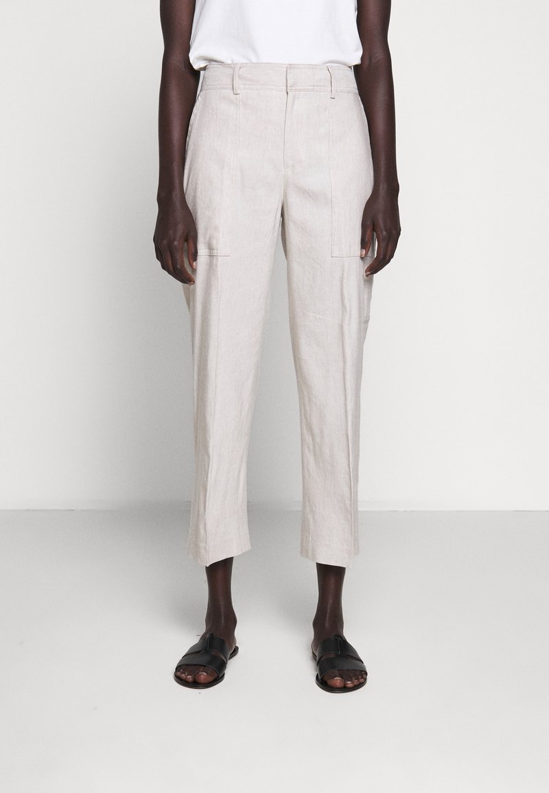 Beige linnebyxor med hög midja, sidofickor och smalnade ben, kombinerat med svarta platta sandaler mot en neutral bakgrund.