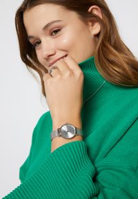 Orologio silver con una cassa rotonda e segna ore sottili, dotato di un cinturino in maglia. Un maglione verde a coste è parzialmente visibile.