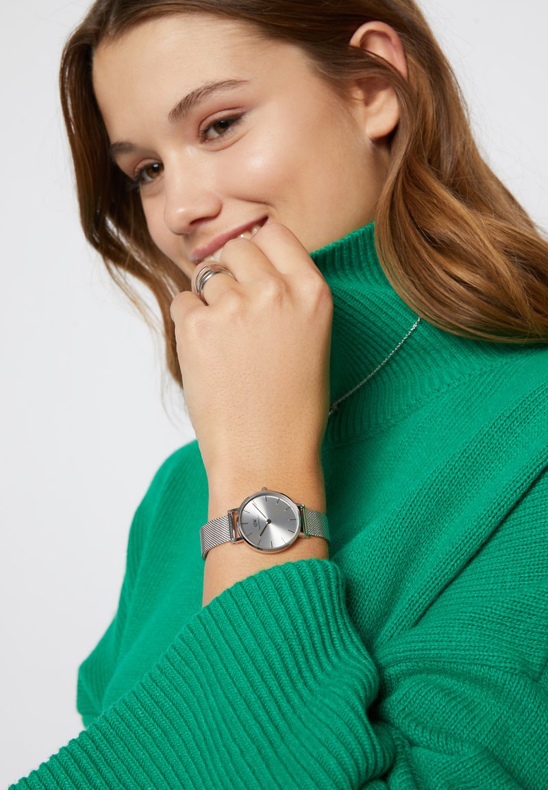 Orologio silver con una cassa rotonda e segna ore sottili, dotato di un cinturino in maglia. Un maglione verde a coste è parzialmente visibile.