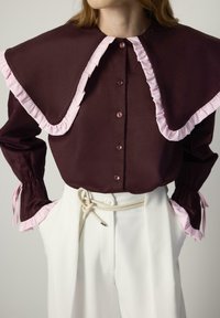 Bordeauxblouse met grote roze gerimpelde kraag, knoopsluiting aan de voorkant en gathered mouwen. Gecombineerd met een witte broek en een touwriem.