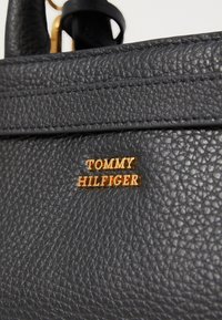 Geantă de mână din piele texturată neagră, cu un logo auriu "TOMMY HILFIGER". Include cusături vizibile și o curea atașată.