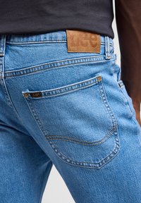 Jasnoniebieskie dżinsy z denimu z skórzaną łatą oznaczoną "Lee" na pasku. Posiadają tylne kieszenie z obszyciami oraz akcenty w kolorze jasnobeżowym.