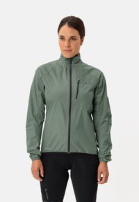 Groene waterdichte fietsjack met een hoge kraag, een rits aan de voorkant, elastische manchetten en een subtiele textuur, gecombineerd met zwarte gevoerde shorts.