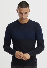 Pulover s pomladnimi modrimi črtami, izdelan iz teksturiranega pletenega blaga. Ima okroglo izrezana vrat in rebraste manšete, majhna oznaka logotipa pa je pri dnu.