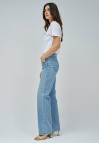 Jeans de tiro alto en azul claro con pierna ancha, hechos de tela de denim. Combinados con una camiseta básica de manga corta blanca y tacones con tiras.