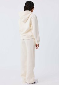 Sudadera color crema y pantalones de chándal a juego, confeccionados en un tejido suave. La sudadera cuenta con un cordón y un bolsillo tipo canguro. Corte holgado.
