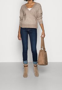 Beżowy sweter typu wrap z mieszanki wełny z dekoltem w serek i ściągaczami przy mankietach, zestawiony z ciemnymi obcisłymi jeansami oraz beżowymi zamszowymi botkami na kostkę. W ręku beżowa torebka.