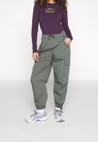 Person in einem lila Levi's California Langarmshirt, lockeren grünen Cargo-Hosen mit Kordelzug und weißen Sneakers mit lila Details.