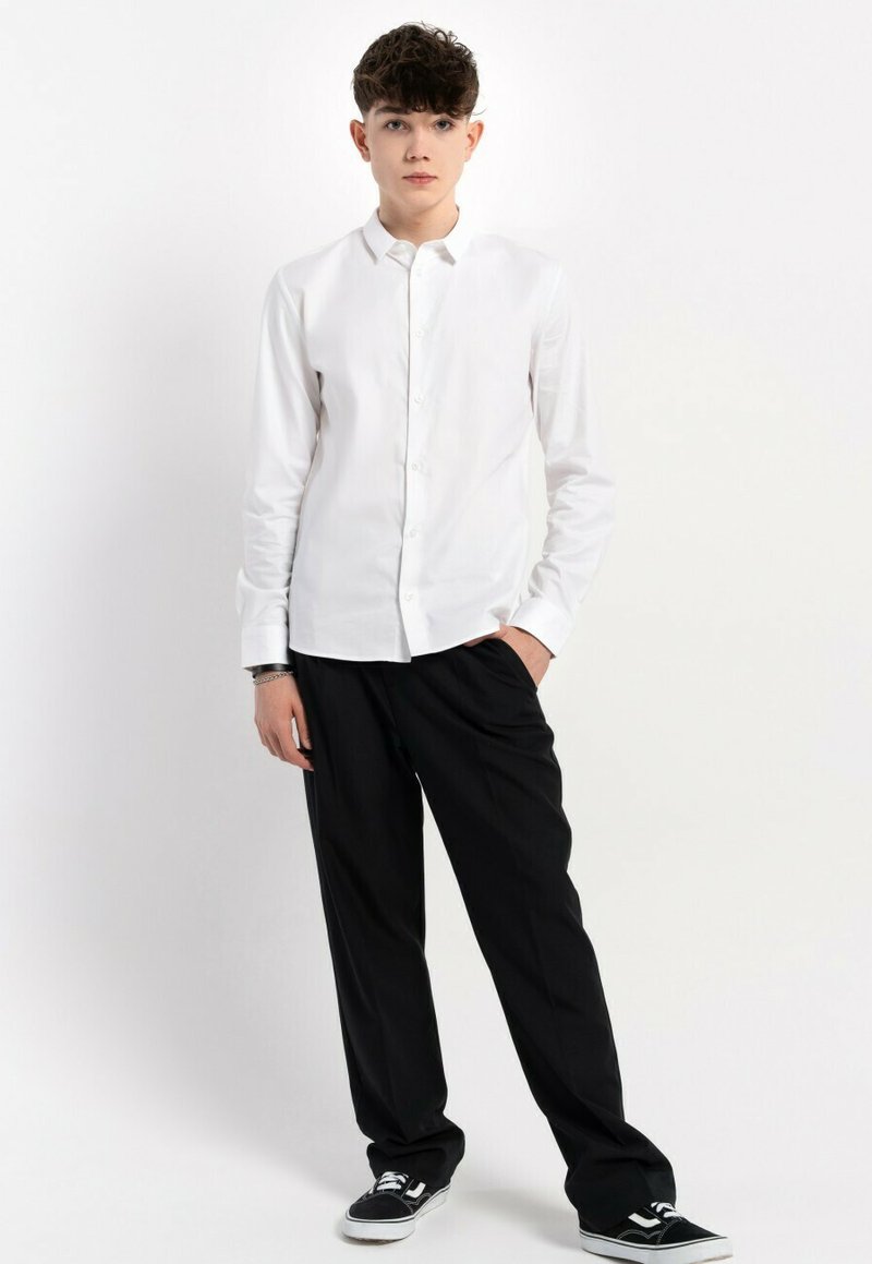 Biała koszula z długim rękawem, zestawiona z luźnymi czarnymi spodniami i czarnymi sneakersami. Koszula ma gładką fakturę i klasyczny krój.