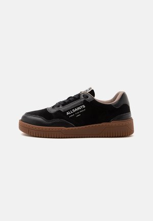 Zapatilla baja negra AllSaints con suela de goma marrón y texto logo blanco en el lateral, mostrada desde el lado exterior.