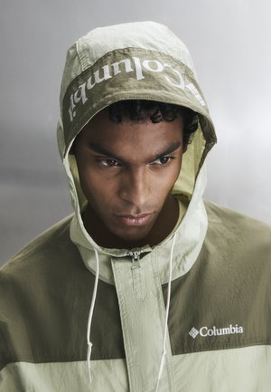 Νεαρός άνδρας με δίχρωμο πράσινο φούτερ Columbia με κουκούλα, κοιτάζει προς τα κάτω σε ουδέτερο φόντο.