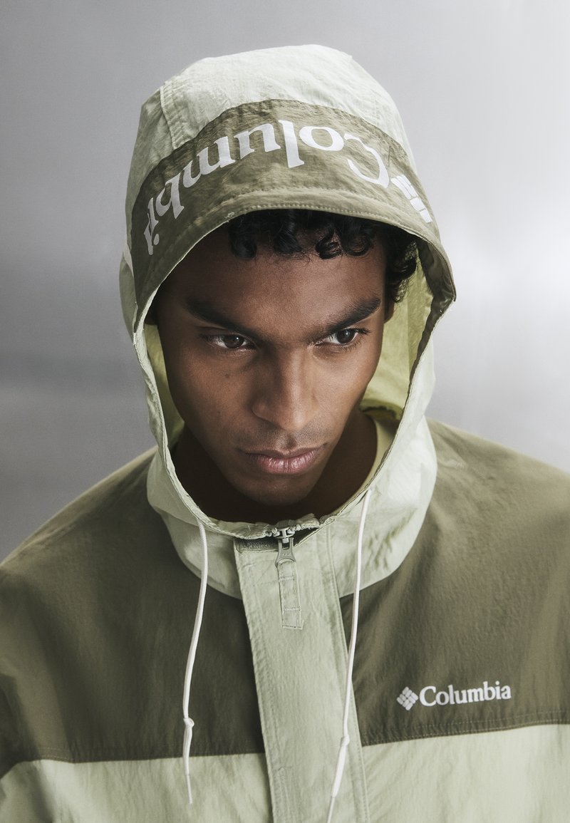 Young man wearing a two-tone green Columbia hooded jacket with the hood up, looking downward against a neutral background.