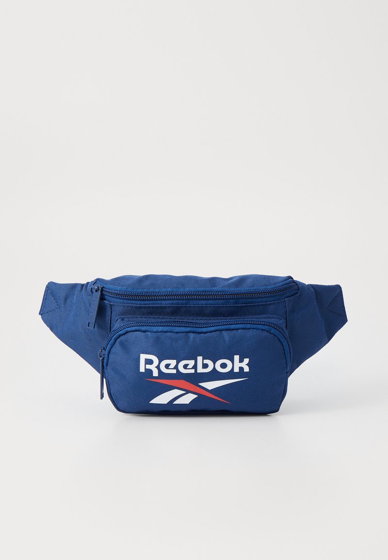 Mochila de cintura em tecido azul marinho com um compartimento principal com fecho e um pequeno bolso frontal com fecho, apresentando um logo da Reebok em branco e detalhes em vermelho.