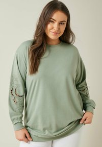 Sudadera de manga larga verde claro, confeccionada en una tela suave. Presenta cortes con costuras decorativas en las mangas. Corte casual con un cuello redondo.