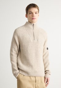 Beige bordás kötött pulóver magas gallérral és fél cipzárral. Jellemzője egy négyzet alakú kivágás a nyaknál és egy logópatch a bal ujjon.