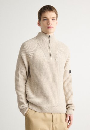 Pull en tricot côtelé beige avec un col haut et une demi-fermeture éclair. Présente une découpe carrée au niveau du cou et une étiquette logo sur la manche gauche.