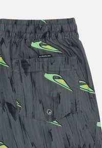 Sivé šortky majú abstraktný vzor v zelenej a žltej farbe, elastický pás a zadnú vrecko so zapínaním na cvoček. Logo je viditeľné na zadnej strane.