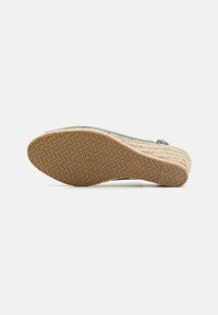 Sandale compensée en espadrille avec une semelle en jute tissée texturée, un dessus en tissu vert clair et une semelle en caoutchouc avec un motif en zigzag.