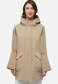 Giacca impermeabile beige con cappuccio, grandi tasche frontali, maniche ampie e chiusura con bottoni automatici. Texture liscia e design minimalista.
