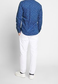 Blå skjorta med knappar och ett litet vitt lövmönster, matchad med vita byxor och vita sneakers, sedd bakifrån.