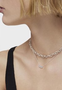 Collares de plata en capas: una cadena gruesa y una cadena delicada con tres pequeñas gemas. El fondo es blanco liso.