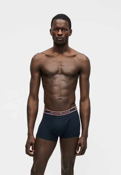 Αθλητικός άνδρας σκουρόχρωμος, όρθιος με το σώμα προς τα εμπρός, φορώντας σκούρο μπλε εσώρουχα boxer Emporio Armani με ροζ ζώνη στη μέση.