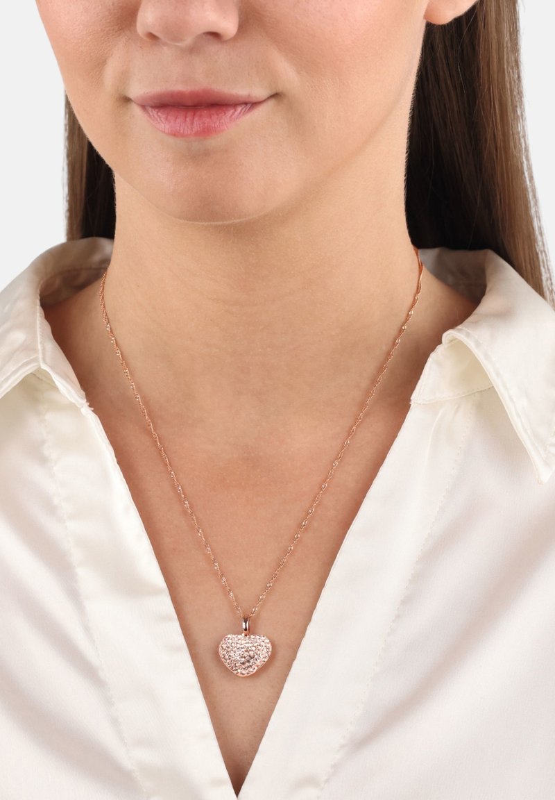 Donna che indossa una collana con pendente a forma di cuore in oro rosa con superficie testurizzata, abbinata a una blusa di raso bianca.