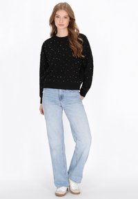 Fekete pulóver, színes pompon díszítésekkel, kerek nyakkivágással, laza fazonban, világoskék egyenes lábú farmernadrággal és fehér sportcipővel párosítva.