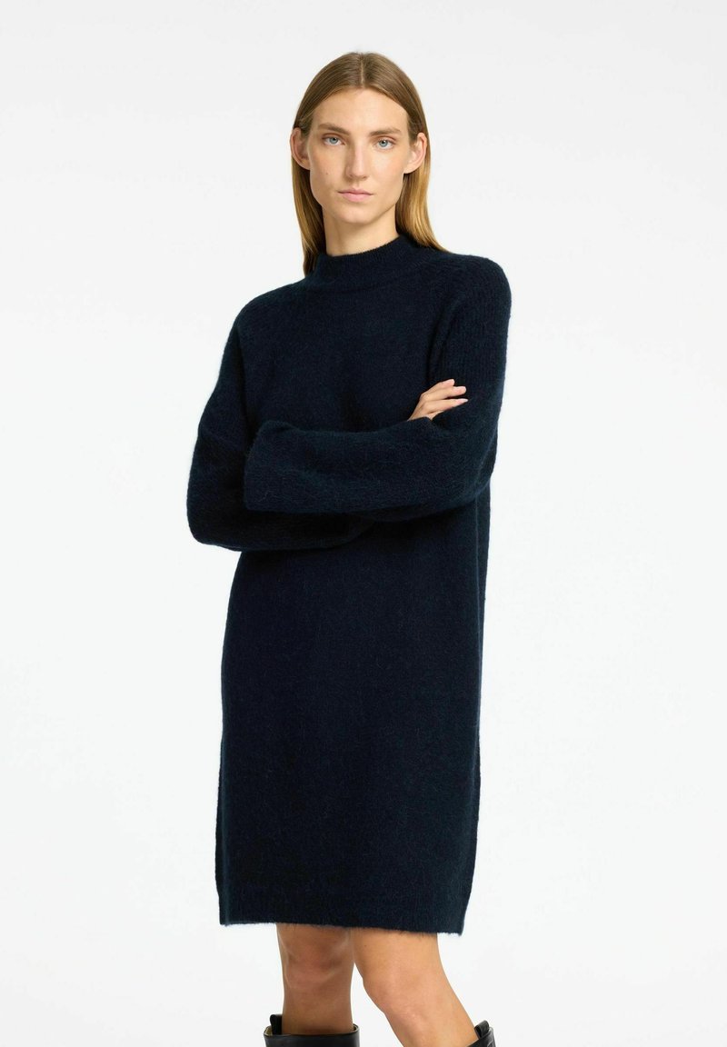 Adīta vilnas kleita tumši zilā krāsā ar augstu kakla izgriezumu, garām piedurknēm un brīvu siluetu. Ievērojamā mīksta, teksturēta apdare.